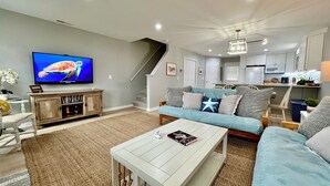 A modern living room with a television, sofa, coffee table, and a staircase leading to a kitchen. - Vast Views | Remodeled Townhome, Pool, Pier Access, Lighthouse Views + Pets (Chincoteague Island)