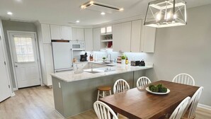 A modern kitchen with white cabinetry, a central island, and a dining area with a wooden table and white chairs. - Vast Views | Remodeled Townhome, Pool, Pier Access, Lighthouse Views + Pets (Chincoteague Island)