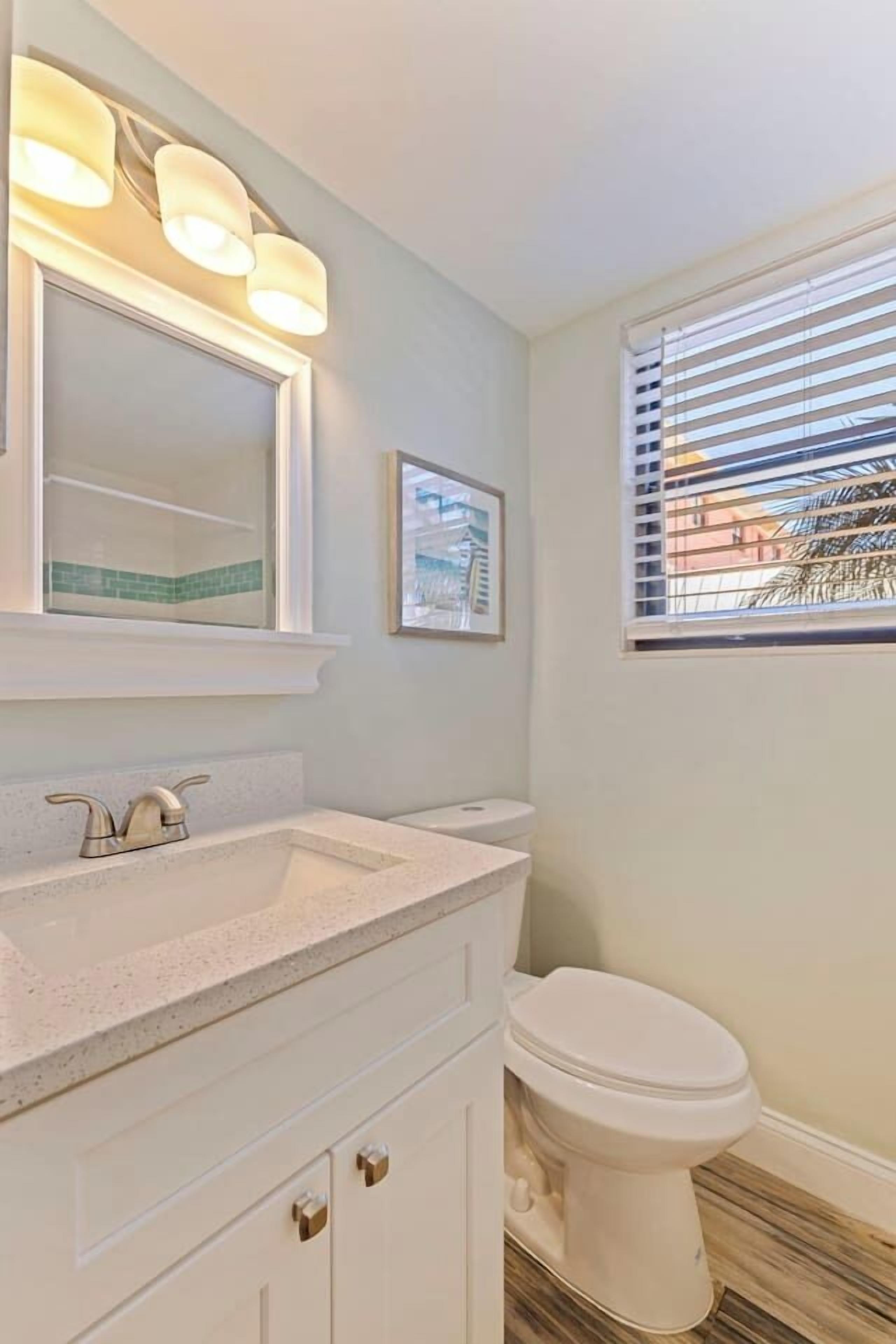 Een badkamer met een wit wastafelmeubel, een spiegel, een toilet en een raam met zonwering.