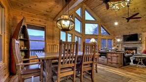 A rustic cabin interior with a wooden dining table, chairs, a fireplace, and large windows. - Mountain Dream Hideaway | Stylish Cabin w/ Mountain Views, Hot Tub & Game Room (Blue Ridge)