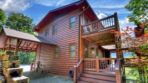 A two-story log cabin with a balcony, a covered porch, and a stone staircase. - Mountain Dream Hideaway | Stylish Cabin w/ Mountain Views, Hot Tub & Game Room (Blue Ridge)