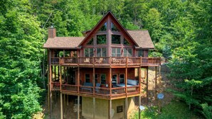 A multi-level log cabin with a balcony, surrounded by dense forest. - Mountain Dream Hideaway | Stylish Cabin w/ Mountain Views, Hot Tub & Game Room (Blue Ridge)