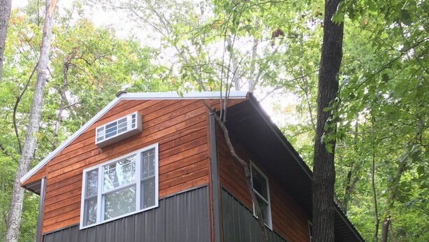 A wooden cabin with a metal roof, built on stilts in a forested area.