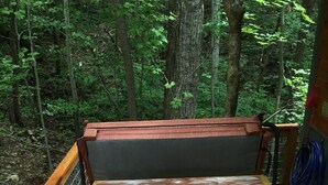 A hot tub with a control panel, surrounded by a wooden deck and metal railing, set in a wooded area.