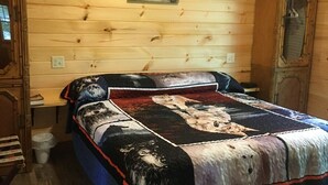 A wooden cabin interior with a bed featuring a wolf-themed quilt, a wall-mounted light fixture, and a wooden cabinet.