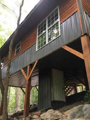 Une cabane à deux étages, dotée d’une terrasse en bois et d’un toit en métal, entourée d’arbres.