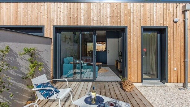 Una casa moderna amb façana de fusta, una porta de vidre que dona a un saló amb un sofà i una taula de menjador preparada amb una ampolla i gots.