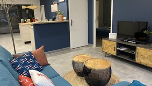 Un saló modern amb un sofà blau, una televisió, taules laterals de fusta i una cuina al fons.