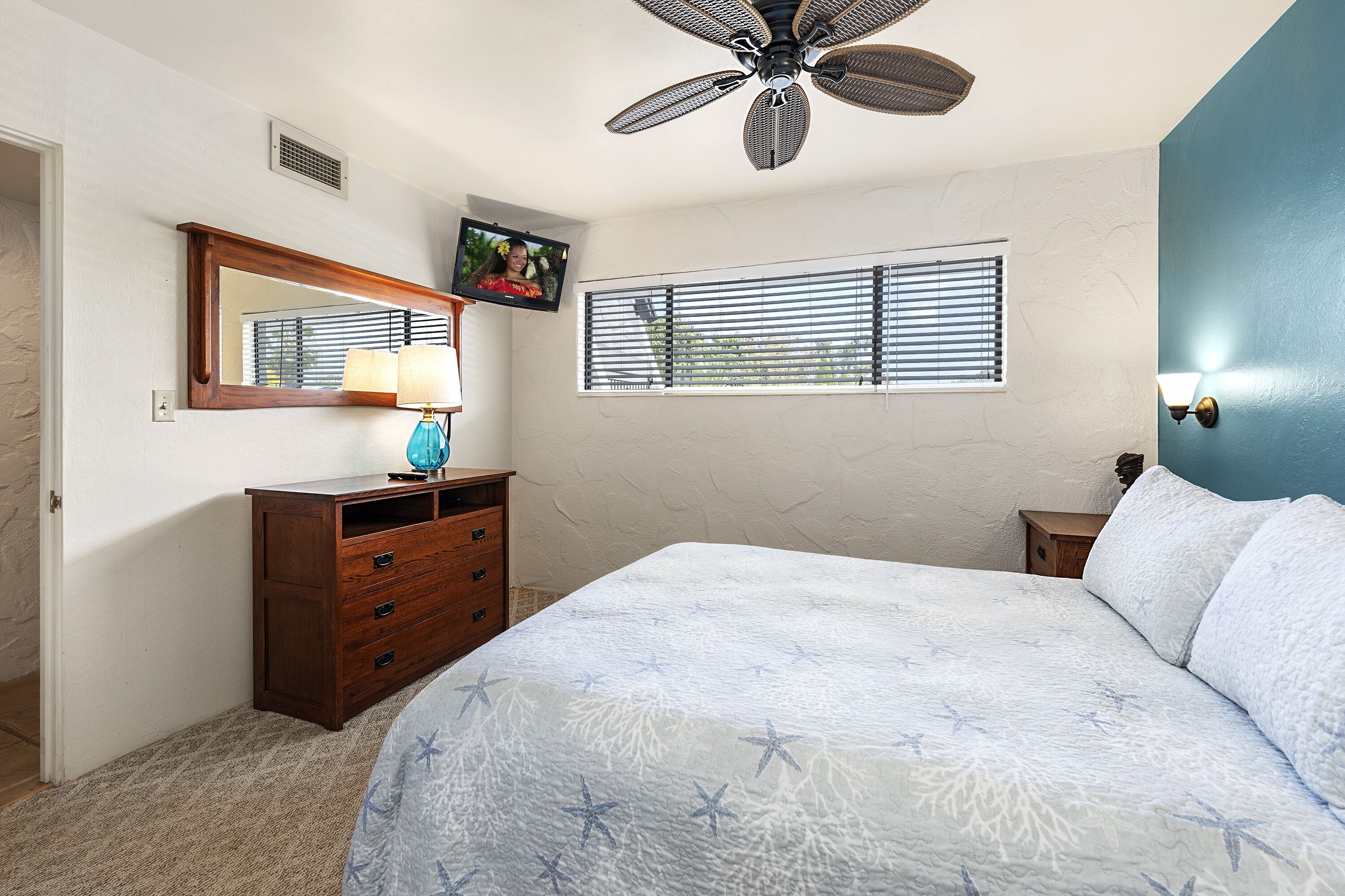 A bedroom with a bed, dresser, mirror, ceiling fan, window, and wall-mounted lamp.