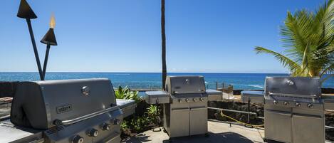 Ein Grillbereich im Freien mit Blick auf das Meer, ausgestattet mit Weber-Grills und Tiki-Fackeln.
