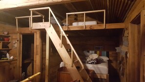 L’intérieur d’une cabane en bois, avec un lit superposé, une échelle en bois et une petite étagère sur laquelle sont posés divers objets.