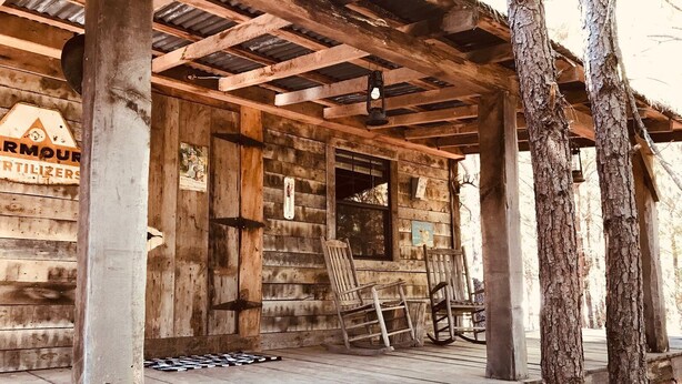 A rustic wooden building with a porch, wooden chairs, and a thermometer on the wall.