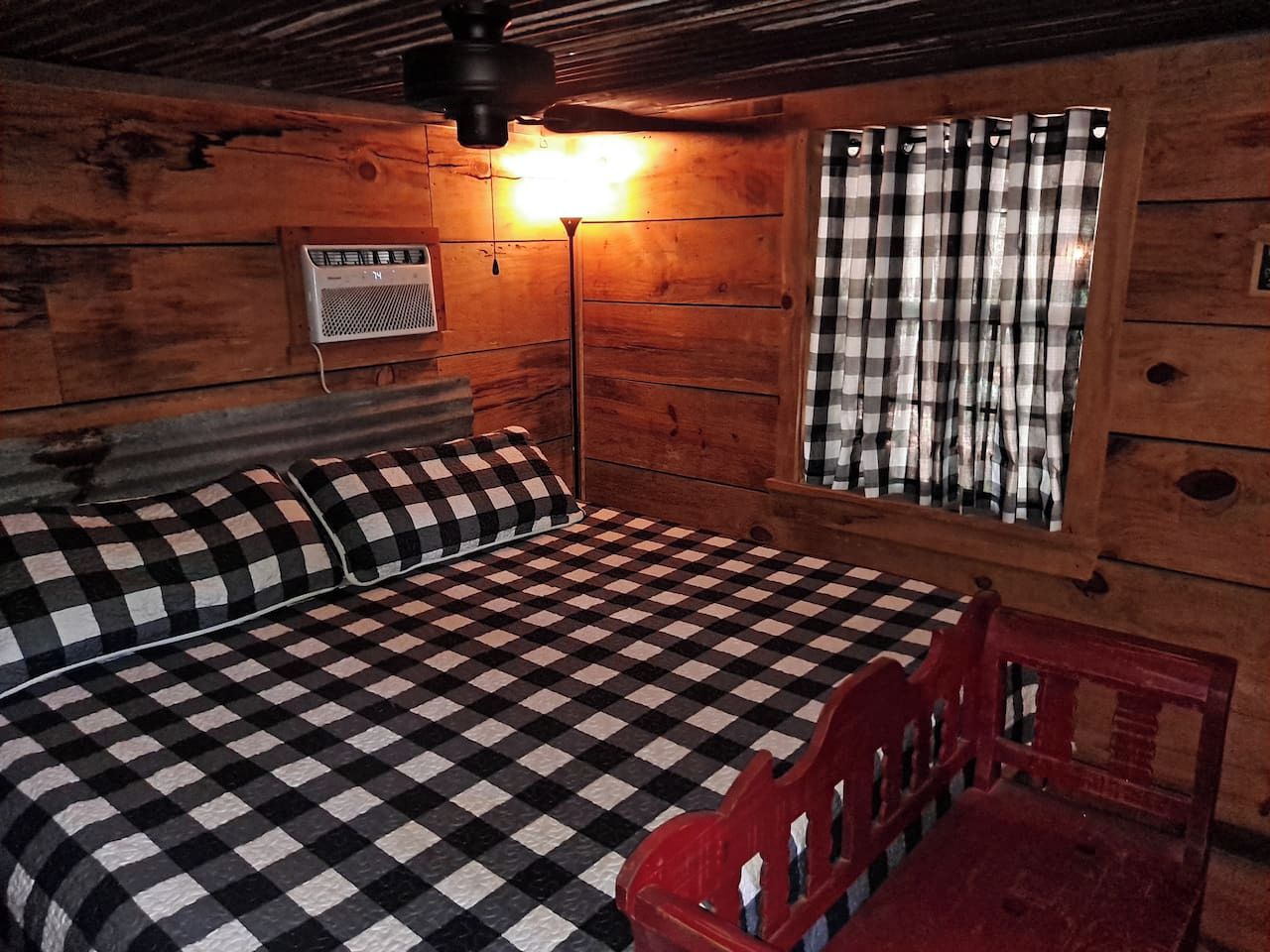 A room with a checkered bedspread, a red chair, a wall-mounted heater, and a window with checkered curtains.