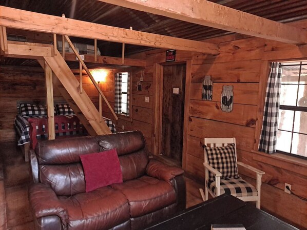 A wooden cabin interior with a leather sofa, a chair, a staircase, and a window with curtains.