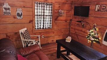 A cozy cabin interior with a wooden floor, a leather couch, a plaid-curtained window, a television, and a small Christmas tree.