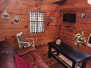 A cozy cabin interior with a wooden floor, a leather couch, a plaid-curtained window, a television, and a small Christmas tree.