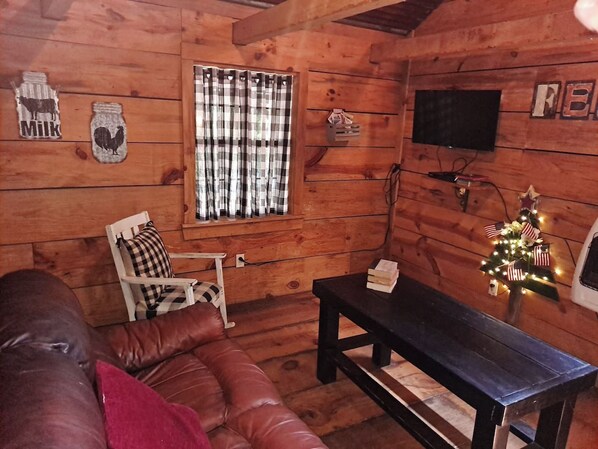 A cozy cabin interior with a wooden floor, a leather couch, a plaid-curtained window, a television, and a small Christmas tree.