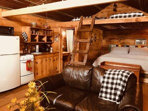 A cozy cabin interior with a kitchenette, a wooden ladder, a bed, and a leather couch.