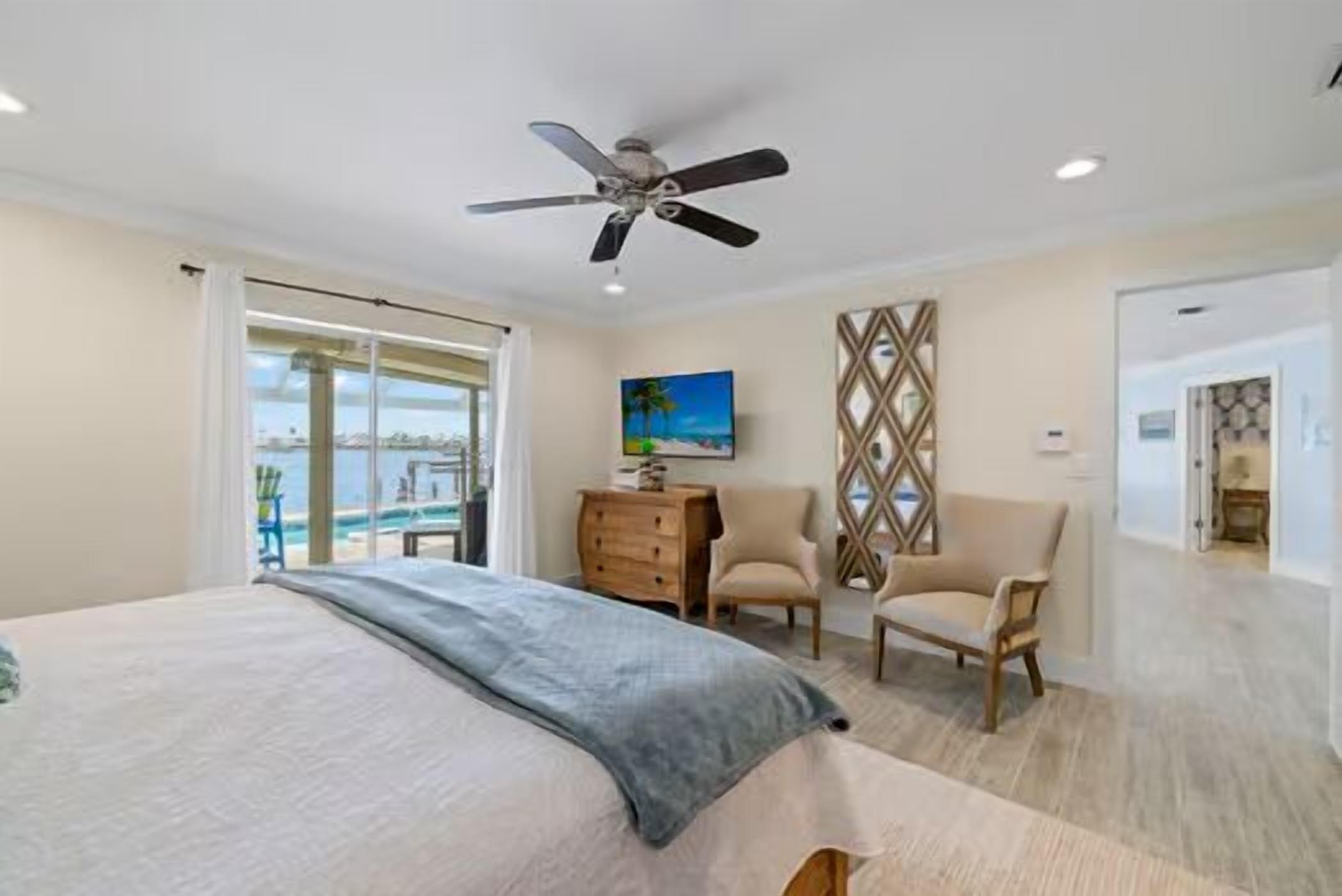 A bedroom with a bed, a ceiling fan, a desk with a chair, a painting, and a view of the outdoors.