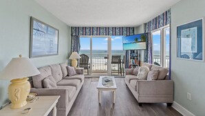 Sala de estar con dos sofás, una mesa de centro y un televisor. Se ve la playa a través de amplias ventanas.