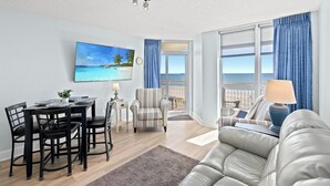 A modern living room with a dining area, a sofa, a lamp, and a painting of a beach scene.