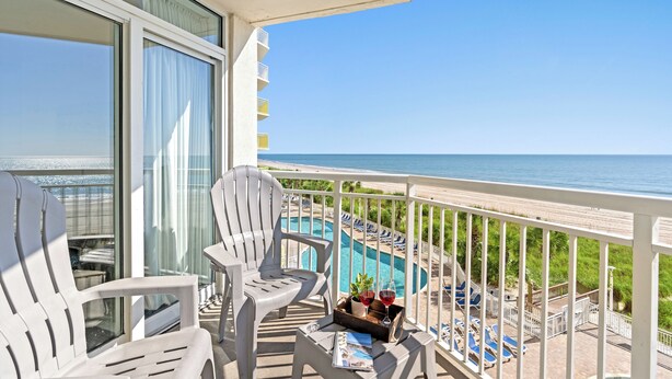 A balcony with two white chairs, a small table with a drink and a plant, and a view of a pool and beach.