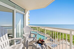 Un balcon avec deux chaises blanches, une petite table avec une boisson et une plante, et une vue sur une piscine et une plage.