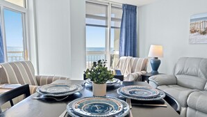 A dining area with a table set for four, a potted plant, and a view of the ocean through large windows.