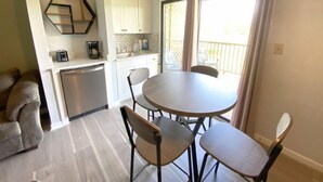 A compact kitchen with white cabinets, a dishwasher, and a coffee maker. There is a round dining table with four chairs. A sofa is visible in the foreground, and a balcony with curtains is seen through the window. - Apple Valley  63 at Rumbling Bald (Lake Lure)