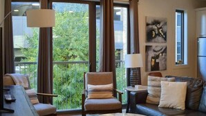 A modern living room with a sofa, armchair, and coffee table, large windows with curtains, and a view of greenery. - Perfect Northern Michigan Escape | Walkable TC Condo | Capri 212 (Traverse City)