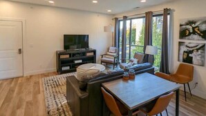 A modern living room with a sofa, a dining table, a TV, and a balcony with curtains. - Perfect Northern Michigan Escape | Walkable TC Condo | Capri 212 (Traverse City)