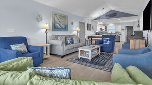 A spacious living room with a sofa, armchairs, and a coffee table. A kitchen is visible in the background. - The Turtle Shell: Home Across from Ocean Crest Pier and Restaurant (Oak Island)
