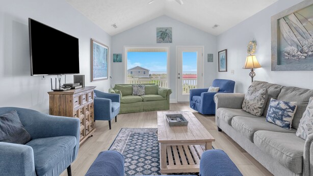 A spacious living room with a large flat-screen TV, a ceiling fan, and a view of a house through a large window. - The Turtle Shell: Home Across from Ocean Crest Pier and Restaurant (Oak Island)