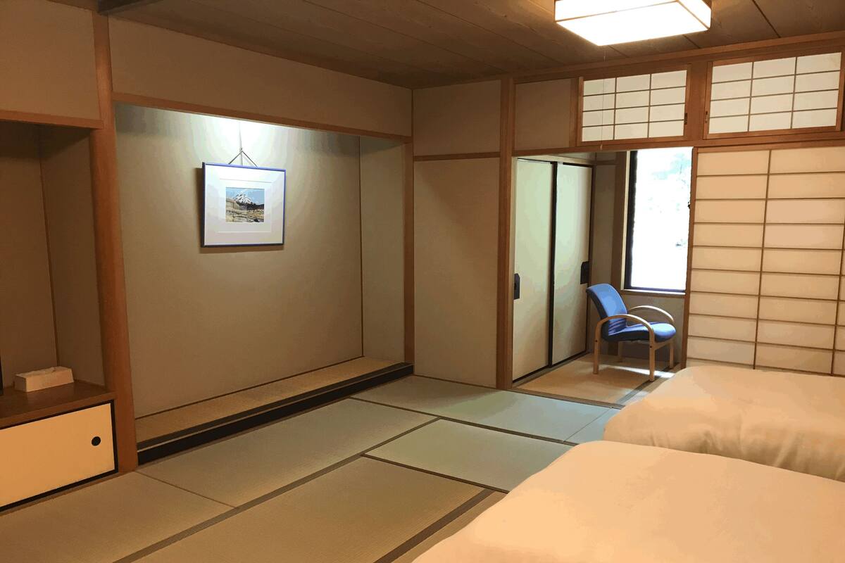 Ein traditioneller japanischer Raum mit Tatami-Böden, einem Bett, einem Sessel und einer Schiebetür.