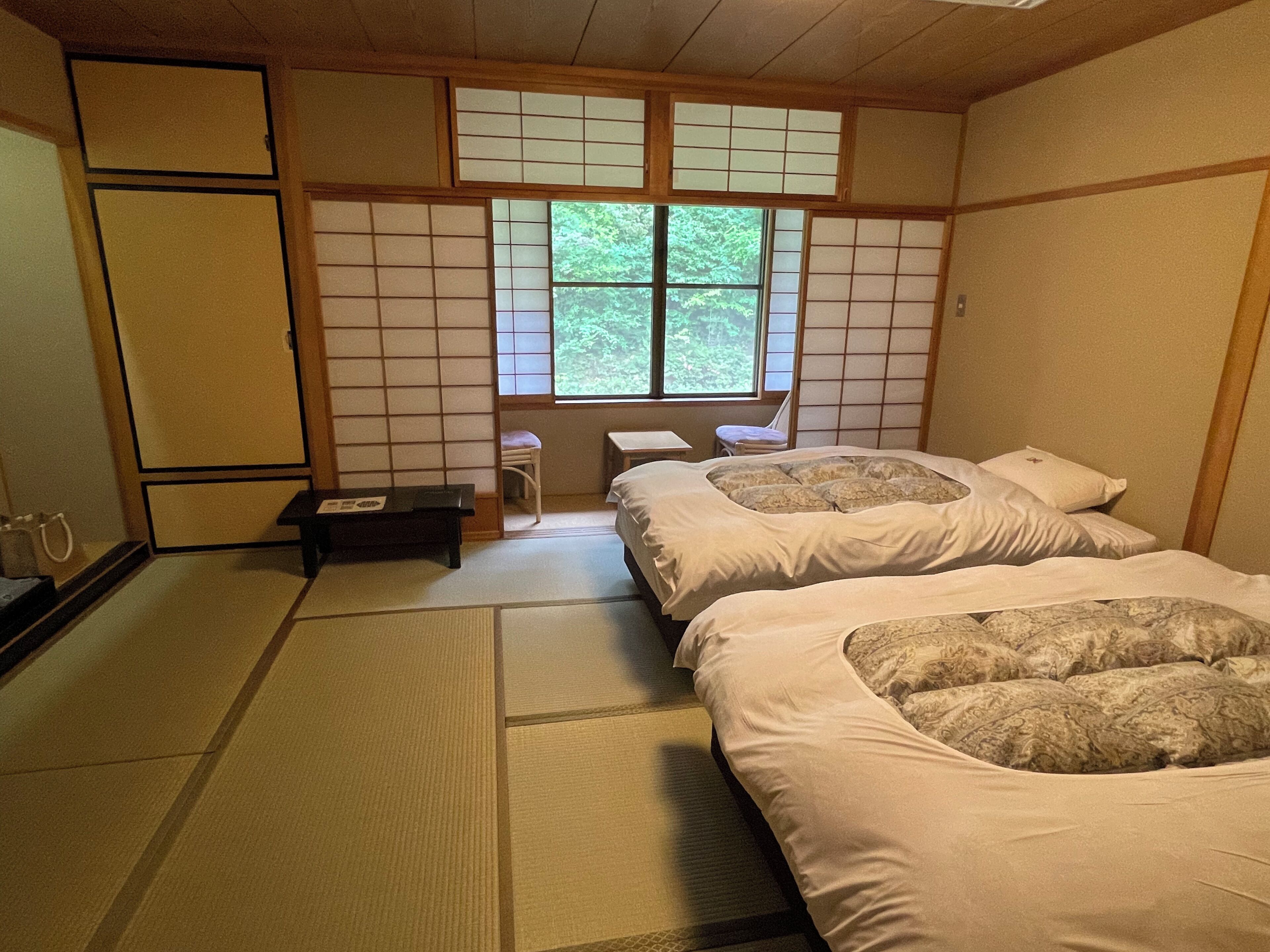 A traditional Japanese-style room with tatami flooring, a futon bed, a small table, and a window with a view of greenery.