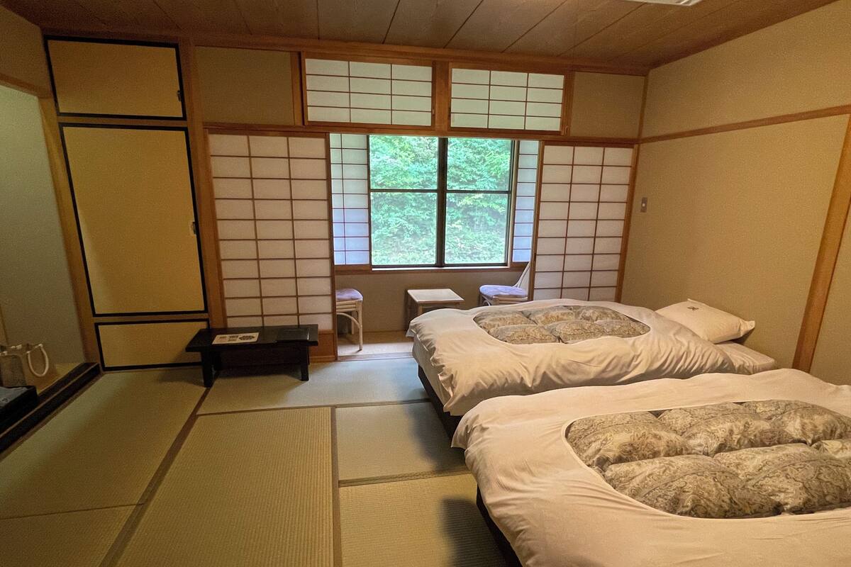 Ein traditioneller japanischer Raum mit Tatami-Böden, einem Futon, einem kleinen Tisch und einem Fenster mit Blick auf Grünflächen.
