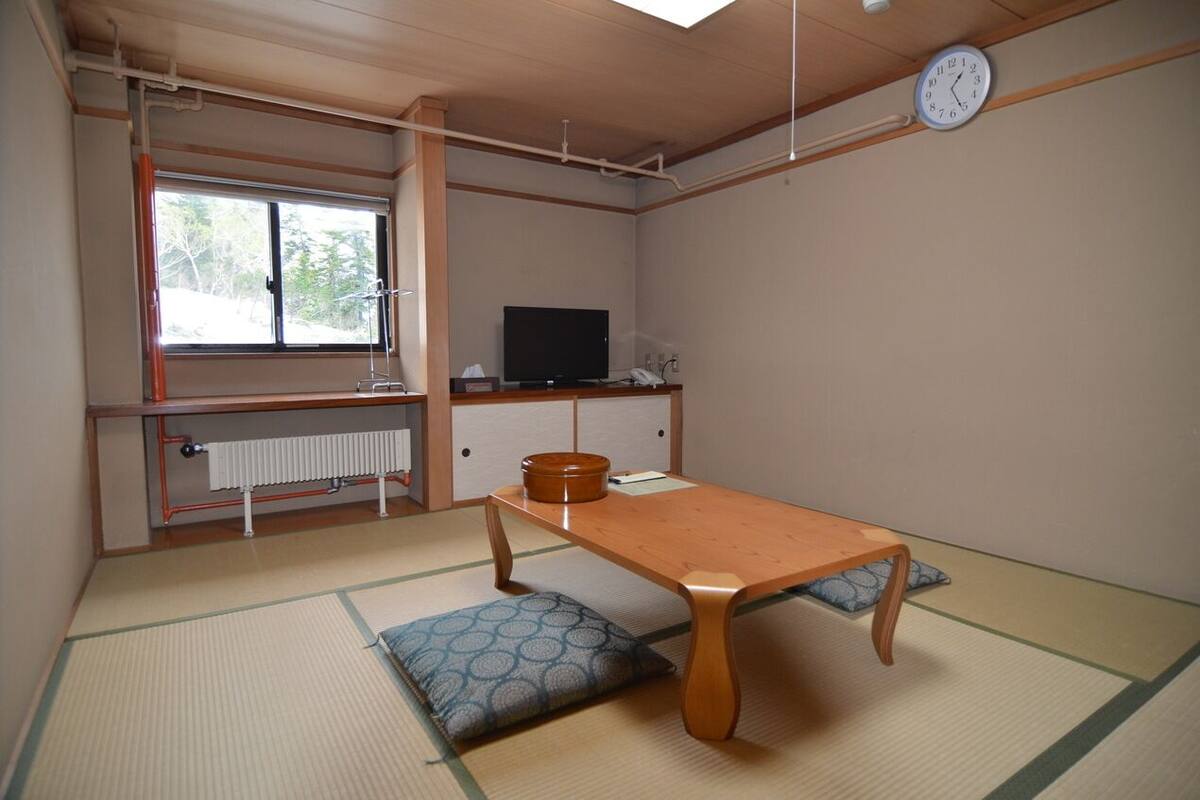 Une pièce de style japonais traditionnel, avec un sol recouvert de tatamis, une table basse, des coussins, un téléviseur et un radiateur.