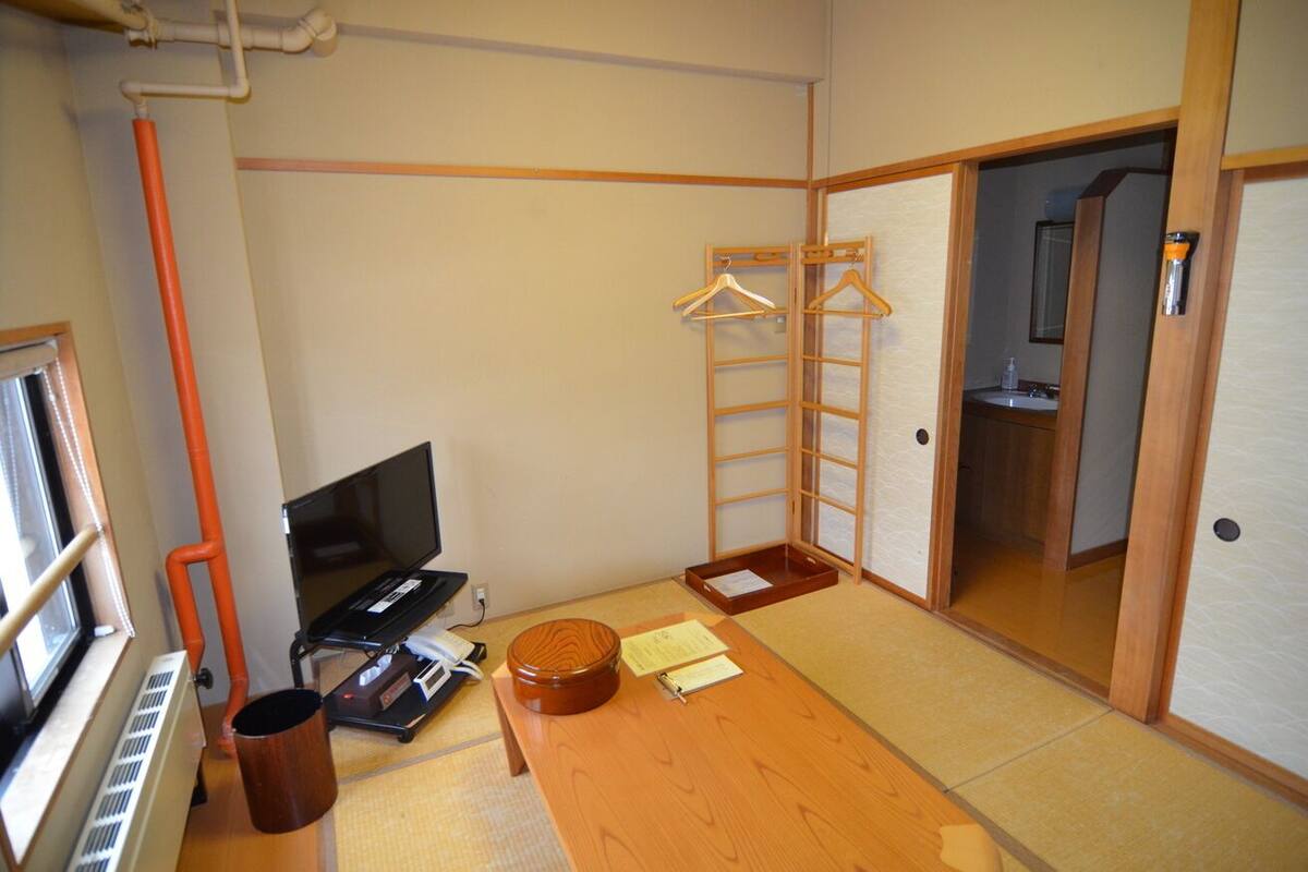 Une pièce de style japonais traditionnel, avec un sol recouvert de tatamis, une table en bois, une télévision, un radiateur, une étagère en bois avec des cintres et une porte coulissante donnant accès à la salle de bain.