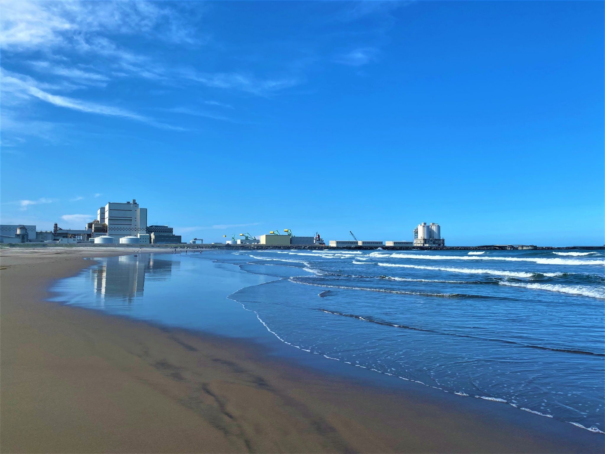 Une plage avec un ciel dégagé, des vagues et, au loin, des bâtiments industriels.
