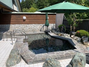 Un jacuzzi con suelo de piedra, una terraza de madera y un paraguas verde.