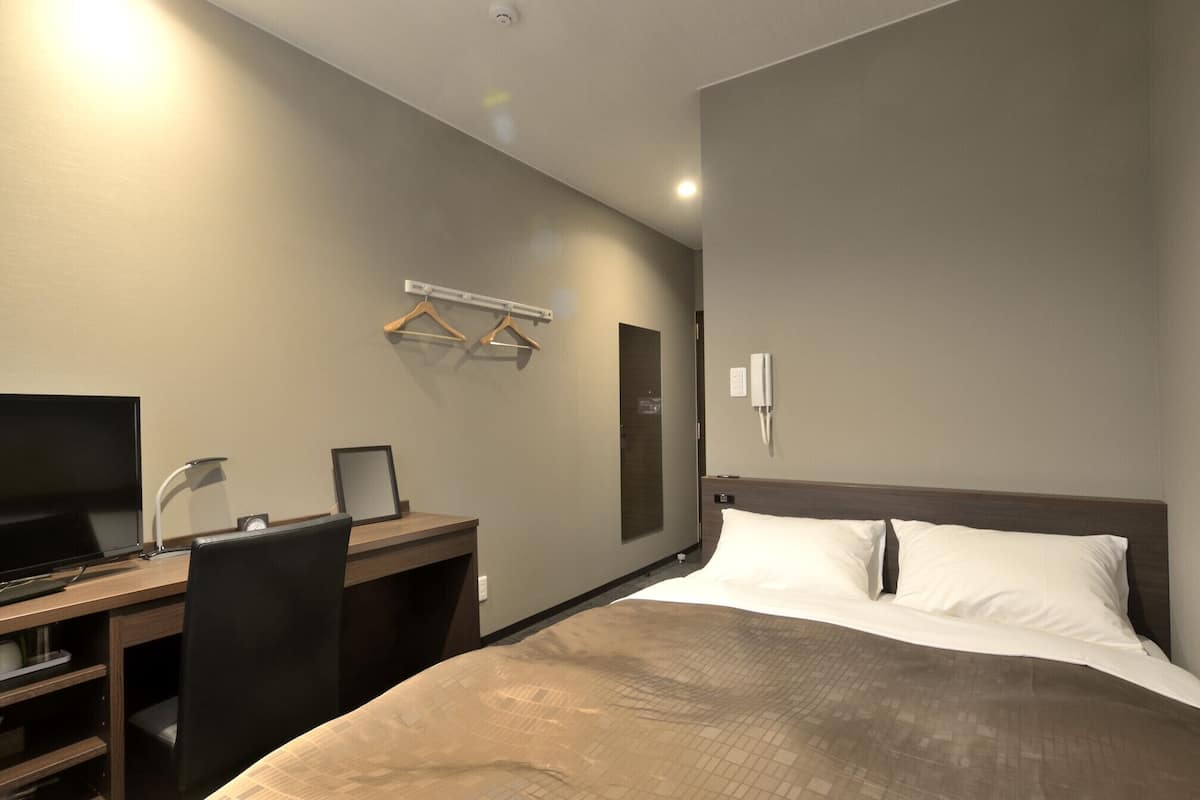 A hotel room with a bed, a desk with a computer, a chair, a TV, and a wall-mounted phone.