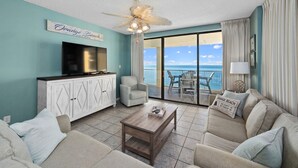 A living room with a television, a ceiling fan, a coffee table, and a sofa. There is a sign on the wall that reads "Orange Beach."