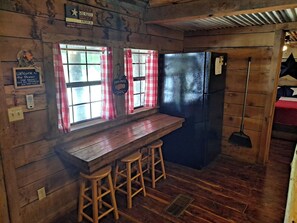 Cocina rústica con barra de madera, dos taburetes, un refrigerador y una ventana con cortinas rojas y blancas.