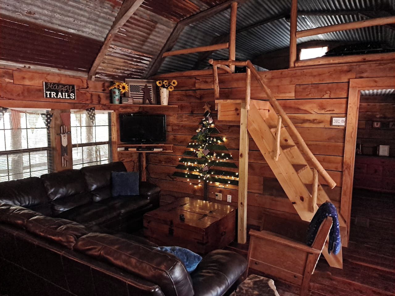 Interior rústico de una cabaña con un sofá de cuero, una escalera de madera, un árbol de Navidad y un letrero que dice 'Happy TRAILS'.