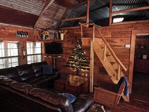 A rustic cabin interior with a leather couch, a wooden staircase, a Christmas tree, and a sign reading 'Happy TRAILS'.