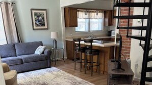 A modern living room with a grey sofa, a coffee table, and a patterned rug. There is a kitchen area with wooden cabinets and a brick wall. - Relax, unwind and enjoy!  (Cody)