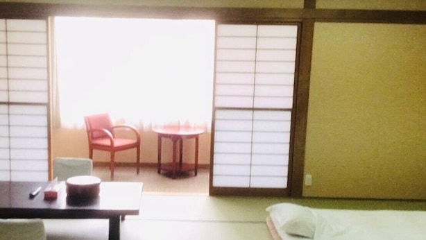 Een traditionele Japanse kamer met tatami vloeren, een laag tafeltje met kussen, een rode stoel en een schuifdeur naar een balkon.