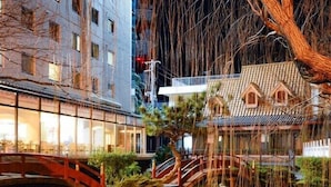 Een traditioneel Japans gebouw met een houten brug over een vijver, omringd door bomen en een modern gebouw op de achtergrond.