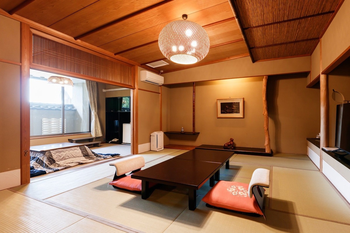 A traditional Japanese-style room with tatami flooring, a low table with cushions, a hanging light fixture, and a television on the right.