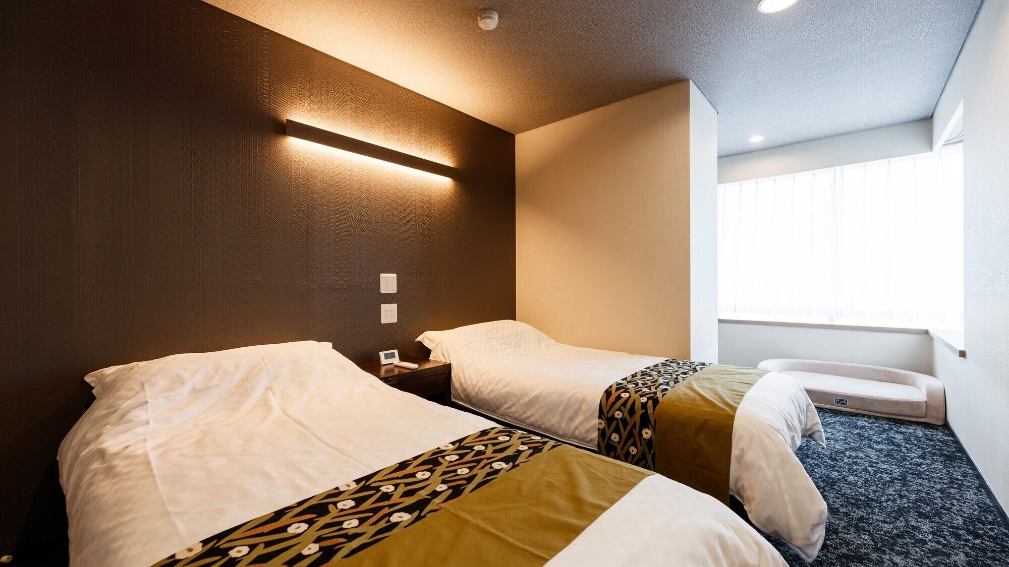A hotel room with two beds, a bedside table with a lamp, a window with blinds, and a carpeted floor.